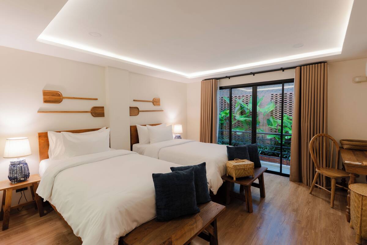 Une chambre d’hôtel avec deux lits, un banc en bois, une table à manger et une vue sur la verdure par la fenêtre.