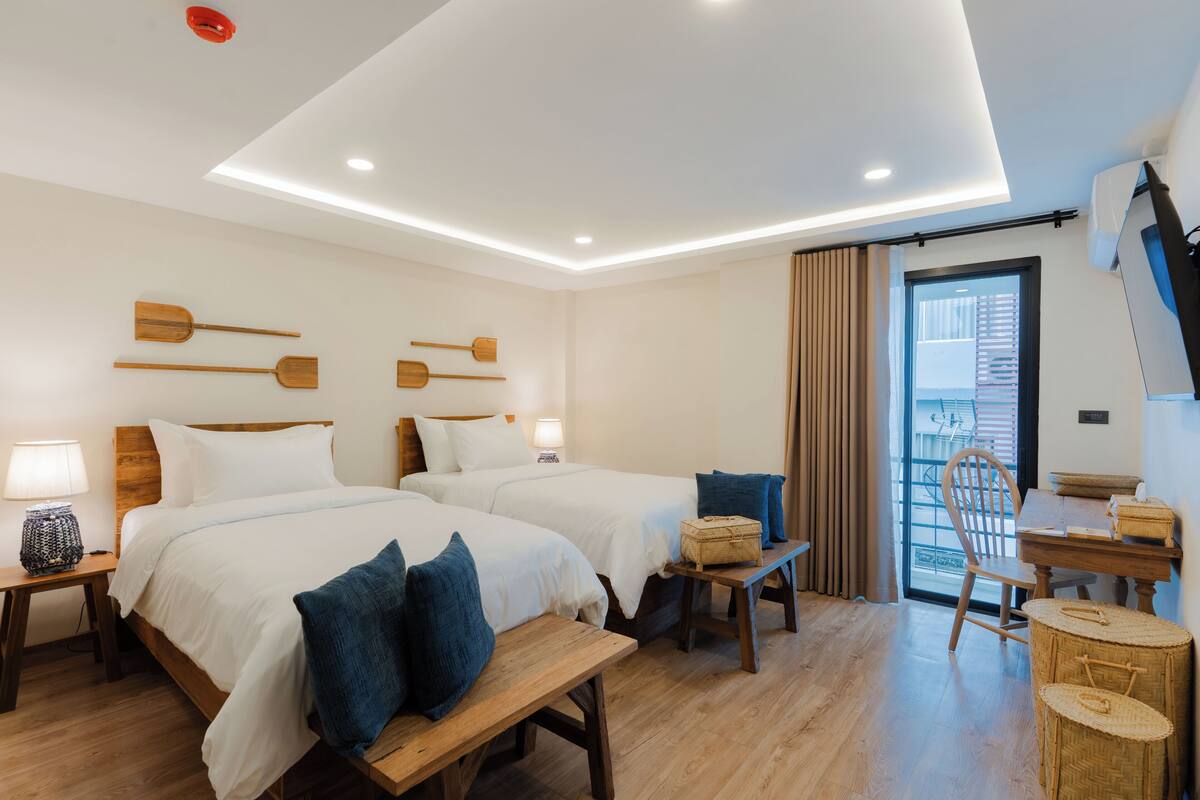 Une chambre d’hôtel avec deux lits, des têtes de lit en bois, une table à manger, une chaise, un banc et une vue sur l’extérieur.