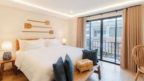 Una habitación de hotel con una cama grande, cabecera de madera, lámparas de noche, un banco de madera, un cofre de mimbre y vistas de la ciudad a través de amplias ventanas.