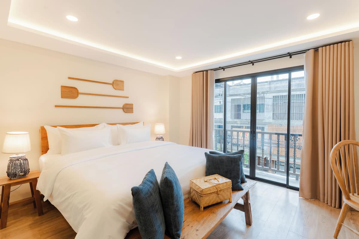 Une chambre d’hôtel dotée d’un grand lit, d’une tête de lit en bois, de lampes de chevet, d’un banc en bois, d’une commode en osier et offrant une vue sur la ville grâce à de grandes fenêtres.
