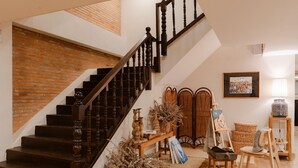 Una escalera con barandillas de madera y un muro de ladrillo. Se ven cestas de mimbre y un cuadro sobre un caballete.