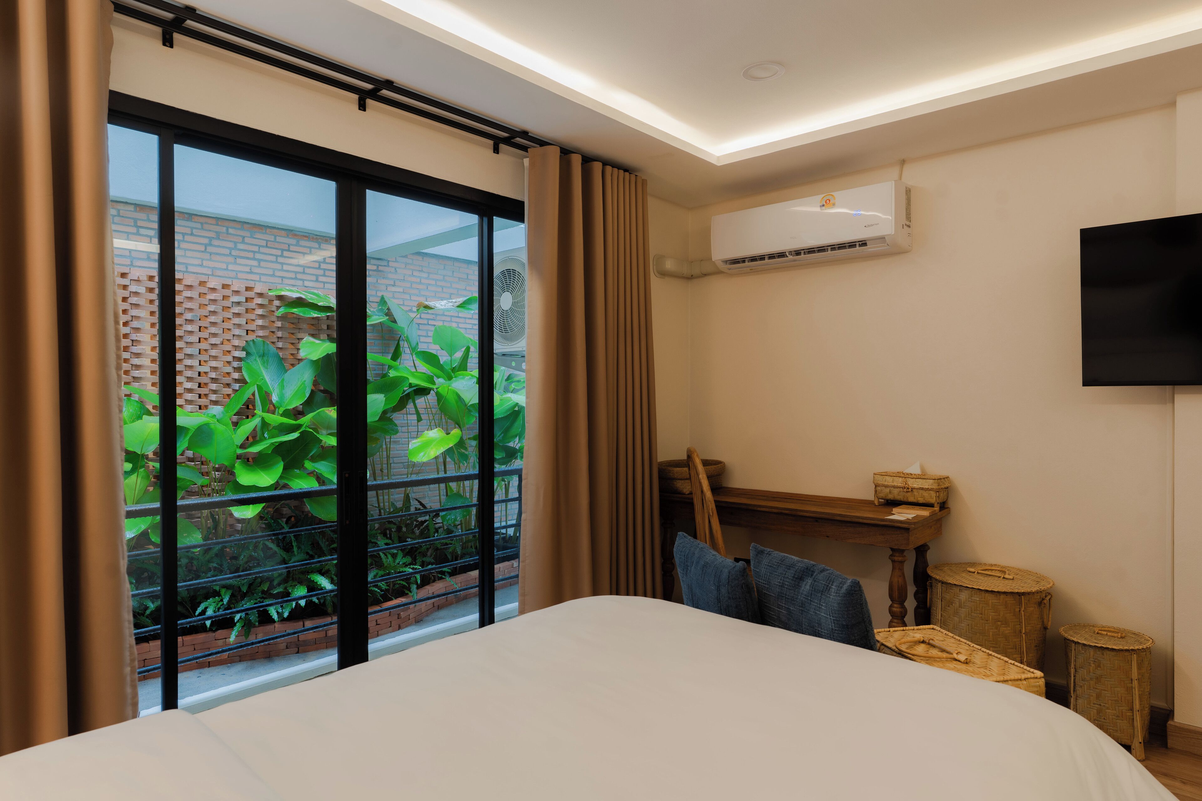 A hotel room with a large sliding glass door leading to a balcony with plants, a flat-screen TV mounted on the wall, and a wooden table with a basket.