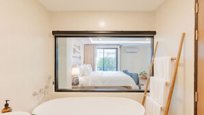 Un baño moderno con una bañera grande, un lavamanos y un espejo que refleja un dormitorio con cama y ventana.