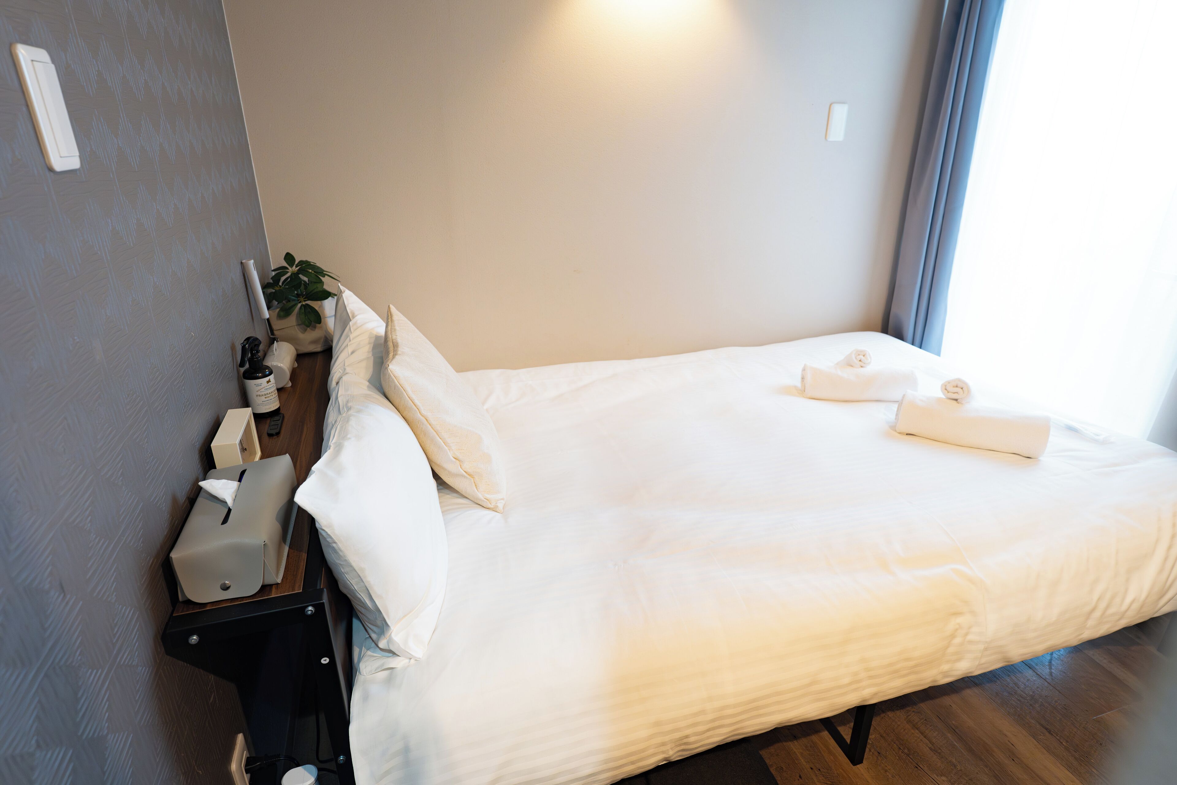 Een opgemaakt bed met witte lakens, een nachtkastje met een lamp, een plant en een flesje lotion.