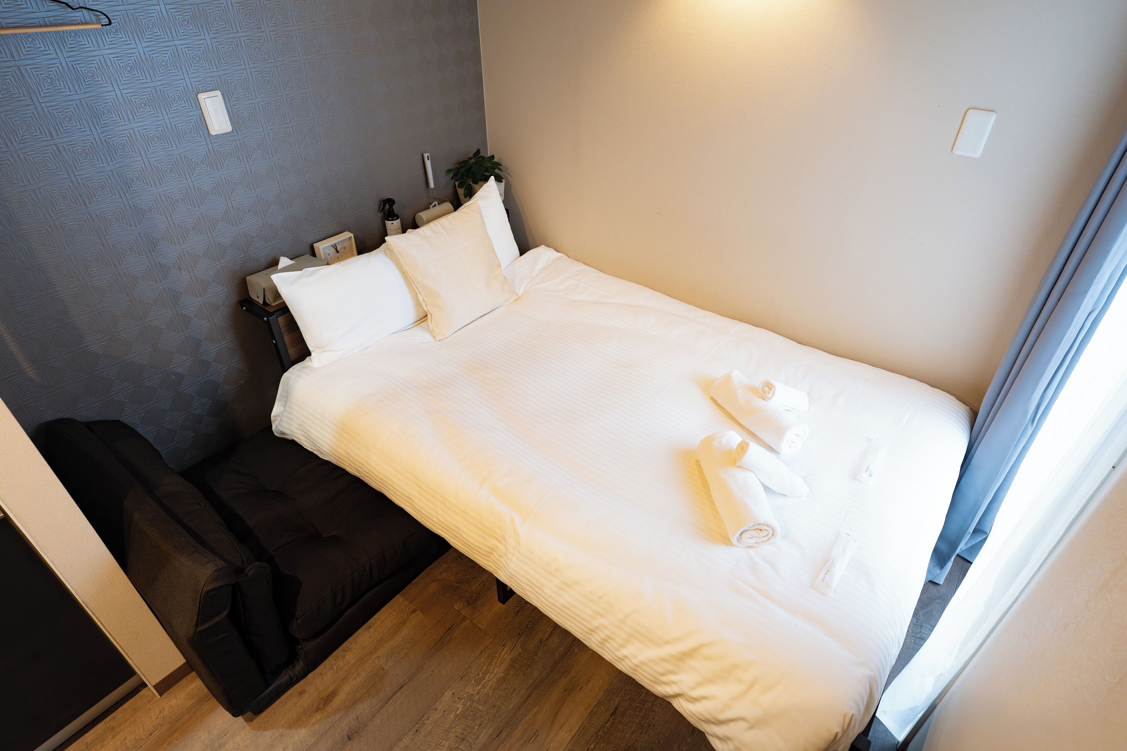 A neatly made bed with white linens and towels, a dark sofa, and a small bedside table with a plant and a bottle.