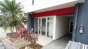 Une entrée d’immeuble avec un auvent rouge, des portes vitrées et un petit jardin planté.