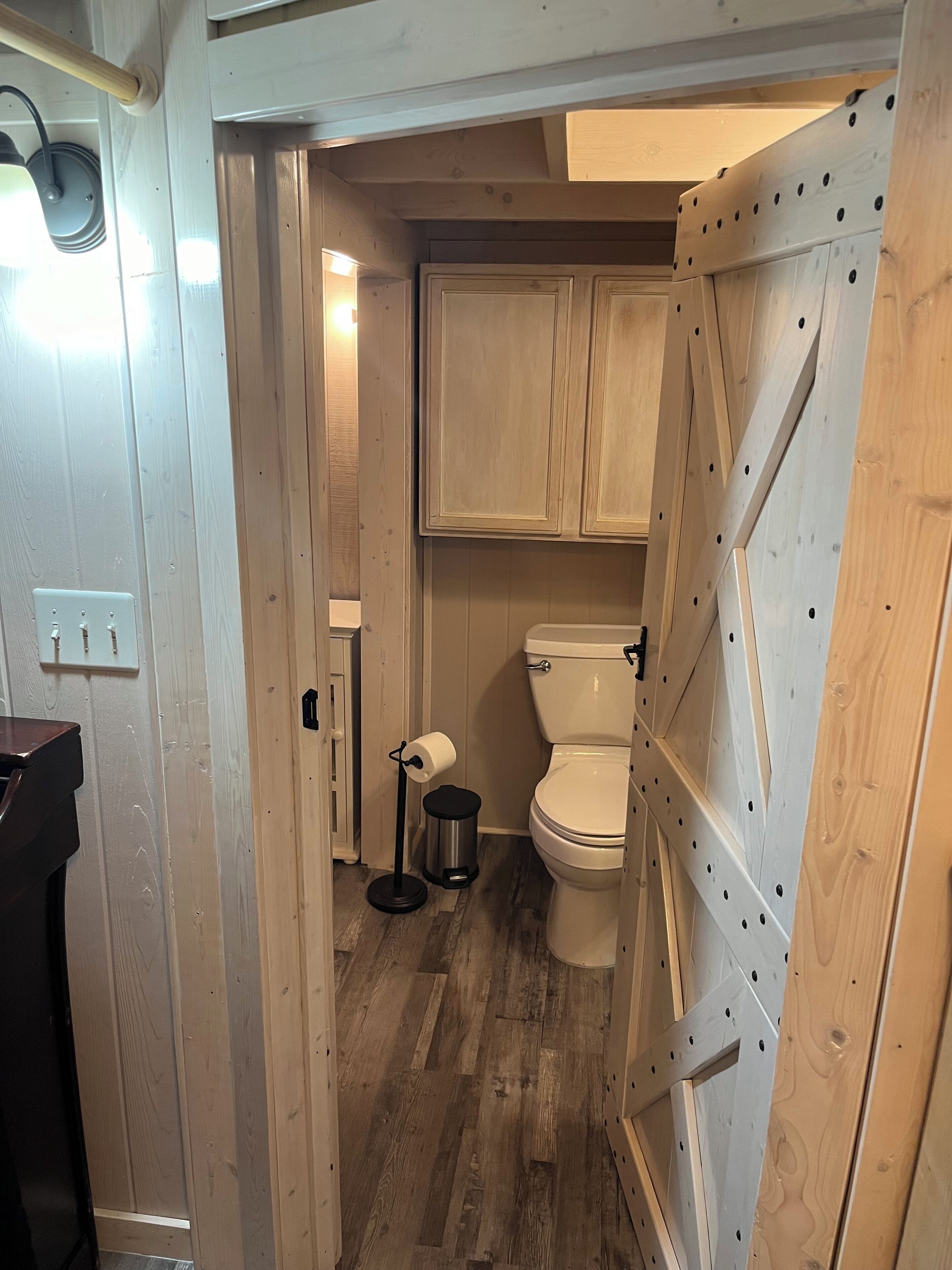 A narrow hallway leading to a bathroom with wooden cabinets, a toilet, and a trash can.