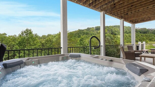 A hot tub with a view of a forested area, a covered patio with outdoor furniture, and a metal railing.