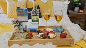 A bed with a book, a bottle, glasses, and a tray with fruits and cheese.