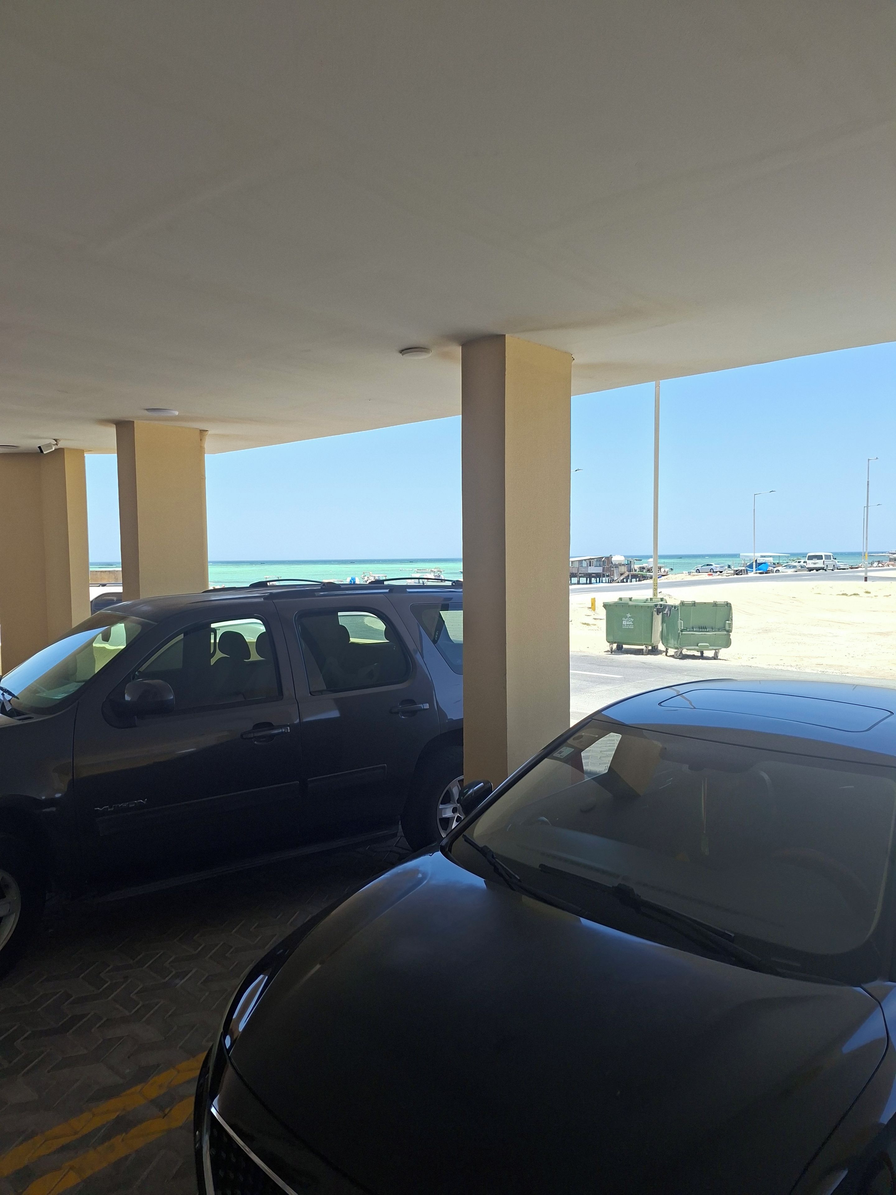 A covered parking area with two cars, a clear view of the beach, and a street with vehicles and dumpsters in the background.