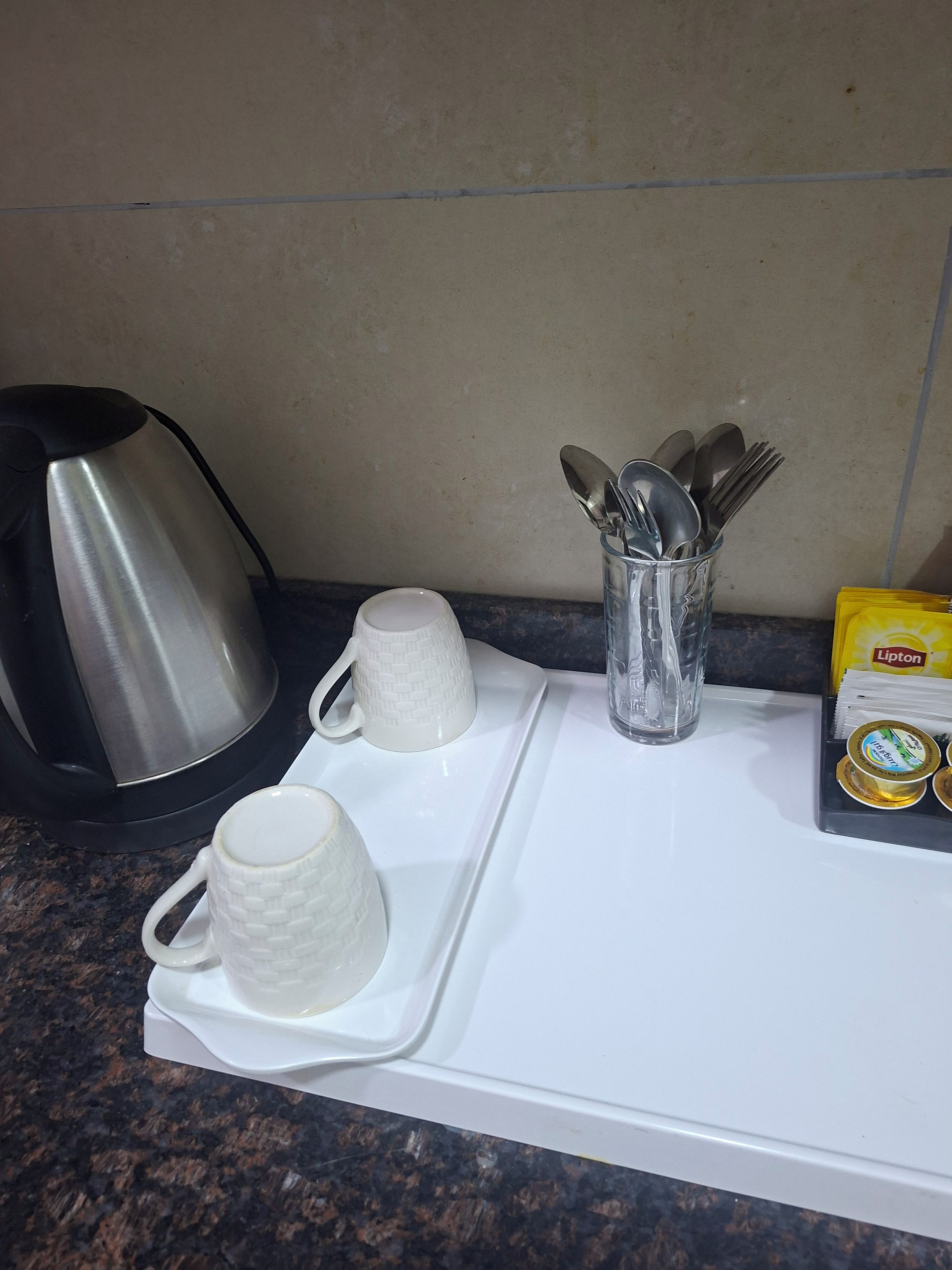 A kitchen counter with a kettle, two white mugs, a glass container with utensils, and a Lipton tea box.