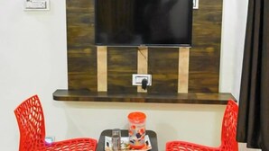 Una stanza modesta con una TV fissata a una parete rivestita in legno, due sedie rosse di plastica, un tavolino con un bicchiere e una bottiglia.