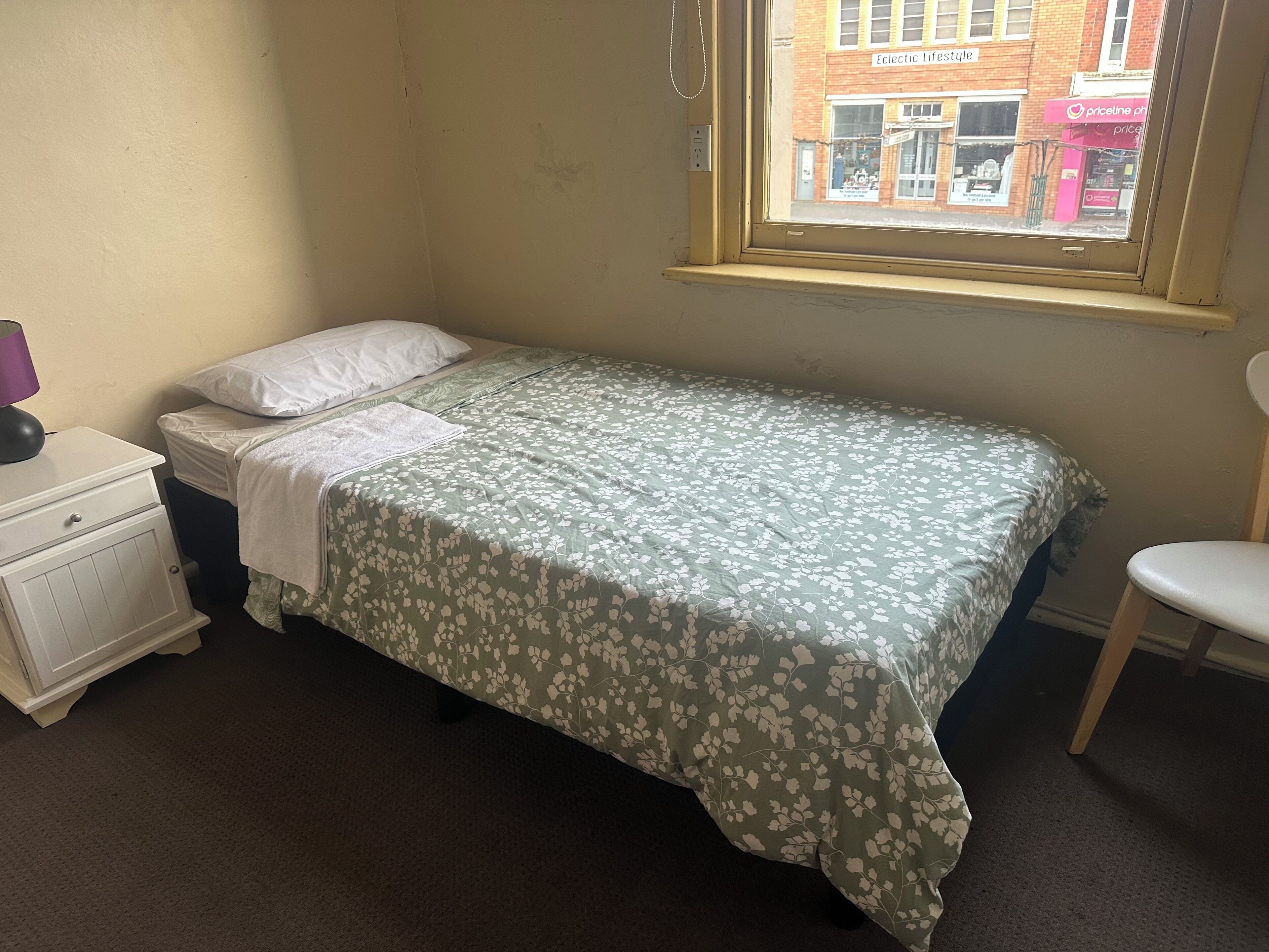 A single bed with a floral bedspread, a white nightstand with a lamp, and a window with a view of a brick building.