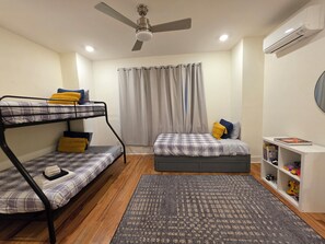 A room with a bunk bed, a single bed, a ceiling fan, an air conditioner, a bookshelf, and a window with curtains.
