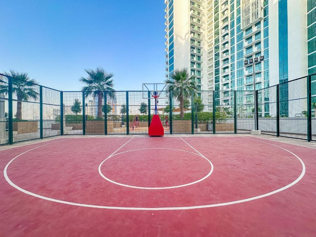An outdoor basketball court with a red basketball hoop, surrounded by a metal fence, and palm trees in the background.