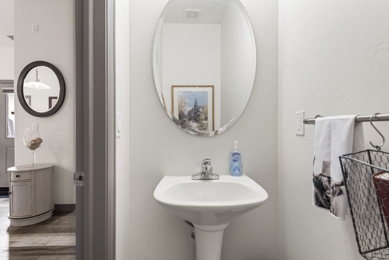 A bathroom with a white sink, a round mirror, a framed picture, a bottle of hand soap, and a towel rack with a towel.