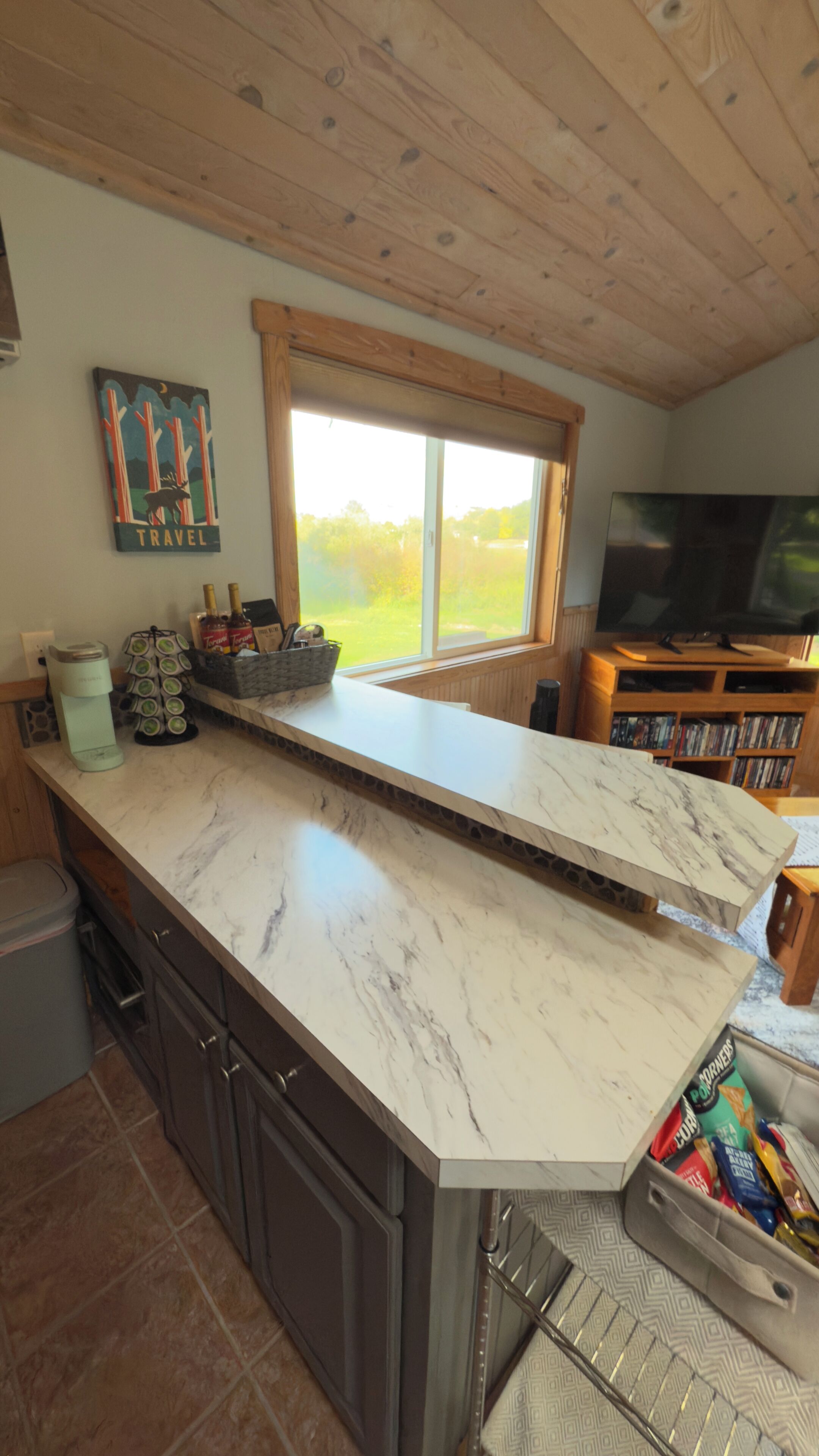 A kitchen with a marble countertop, wooden cabinets, a window with a view, and a television in the background.