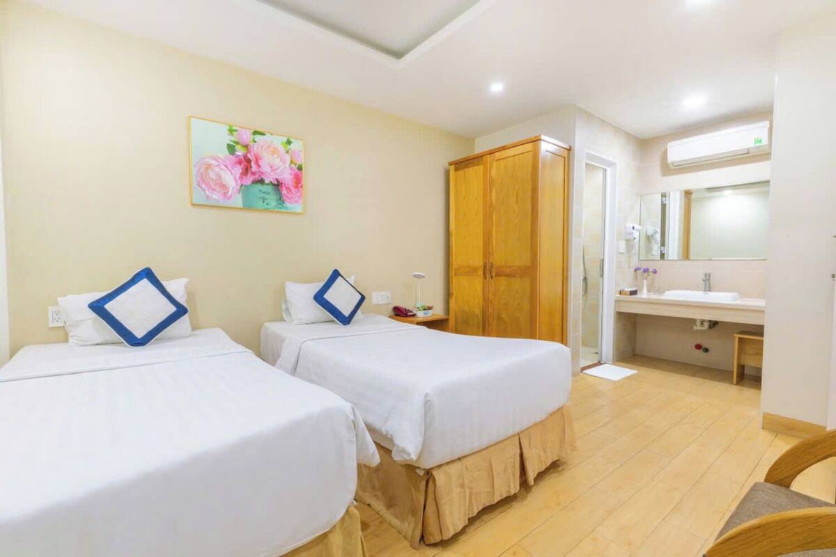 A hotel room with two beds, a wooden wardrobe, a bathroom with a sink and mirror, and a floral painting on the wall.