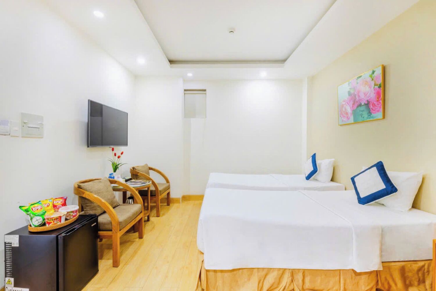 A hotel room with two beds, a TV, a small table with chairs, and a refrigerator.