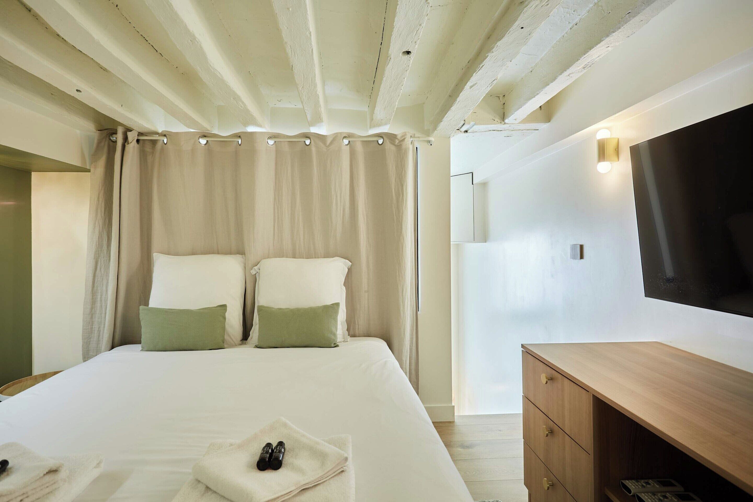 A hotel room with a bed, a wooden dresser, a flat-screen TV, and a curtain.