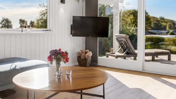 A modern living room with a round wooden table, a grey sofa, a TV mounted on the wall, and a large sliding glass door leading to an outdoor area with a pool and lounge chairs.