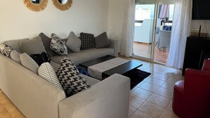 Sala de estar com sofá cinza, almofadas estampadas, mesa de centro preta e uma cadeira vermelha. Há dois espelhos fixados na parede e um lustre no teto.
