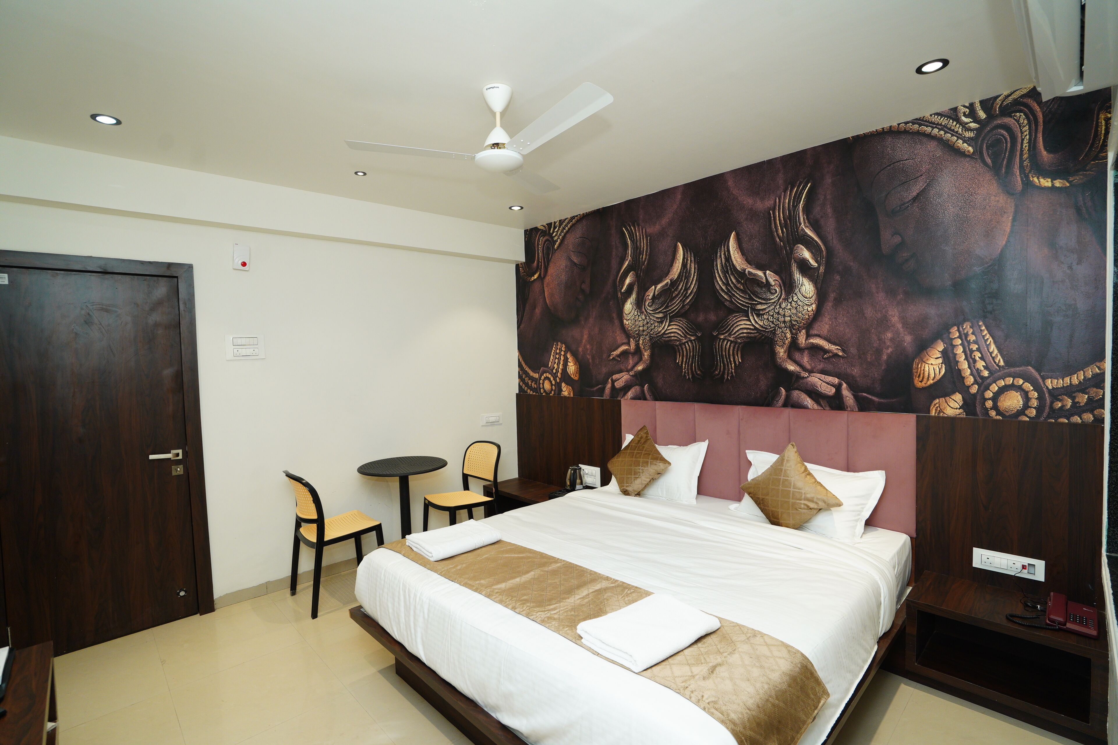 A hotel room with a bed, a chair, a small table, a ceiling fan, and a wall with a decorative mural.