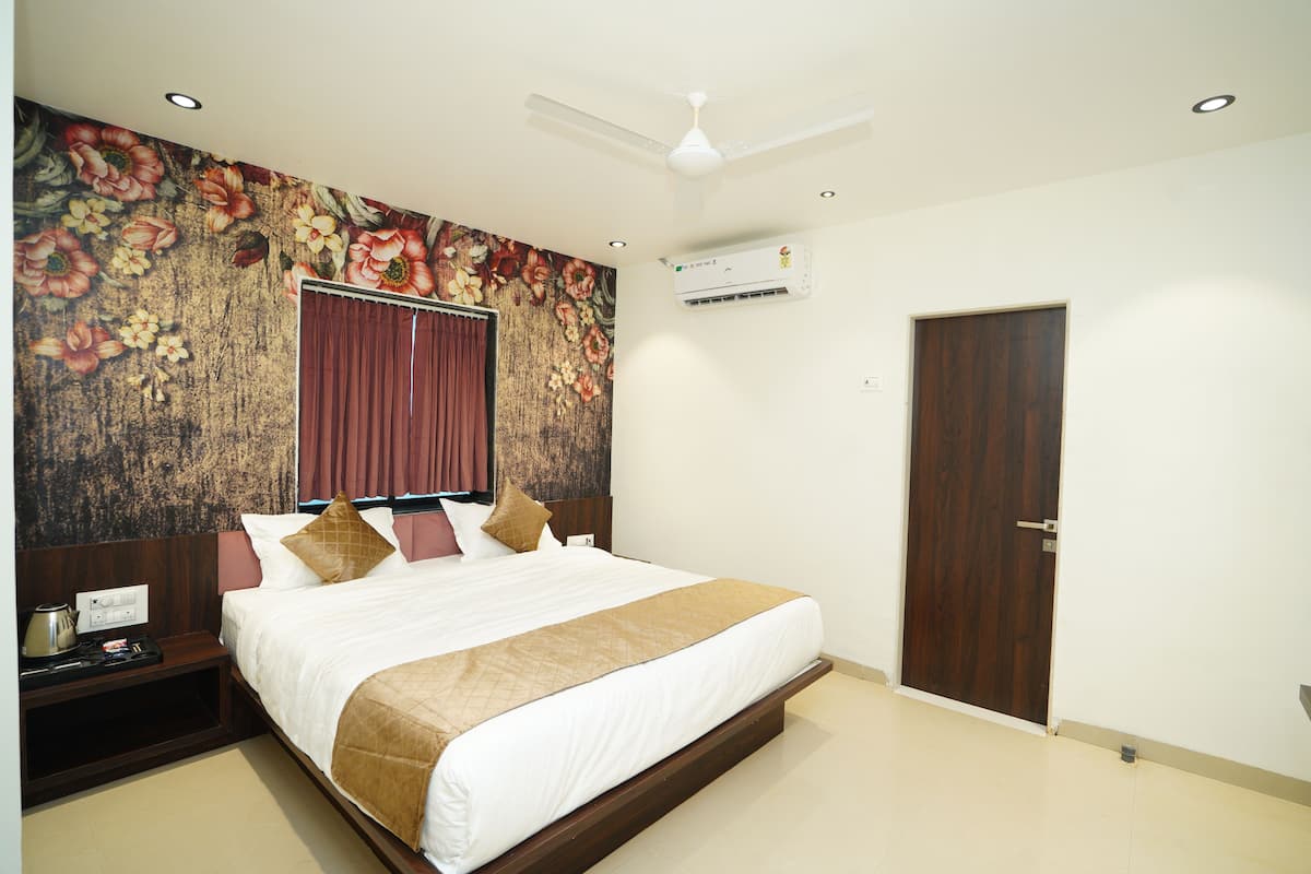 A hotel room with a large bed, a wooden headboard, a floral mural, a ceiling fan, an air conditioning unit, a wooden door, and a nightstand with a kettle and a cup.