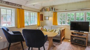 Una habitación acogedora con piso de madera, un sofá, dos sillones, una mesa, un televisor y ventanas grandes con cortinas amarillas.