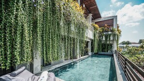 Ein Schwimmbecken mit einer Holzterrasse und Liegestühlen, umgeben von üppiger Vegetation und einem modernen Gebäude im Hintergrund.