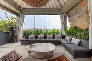 A modern outdoor seating area with a round table, cushioned sofas, and a view of the cityscape through large windows. - Duur Apartments by Betterplace (Seminyak)