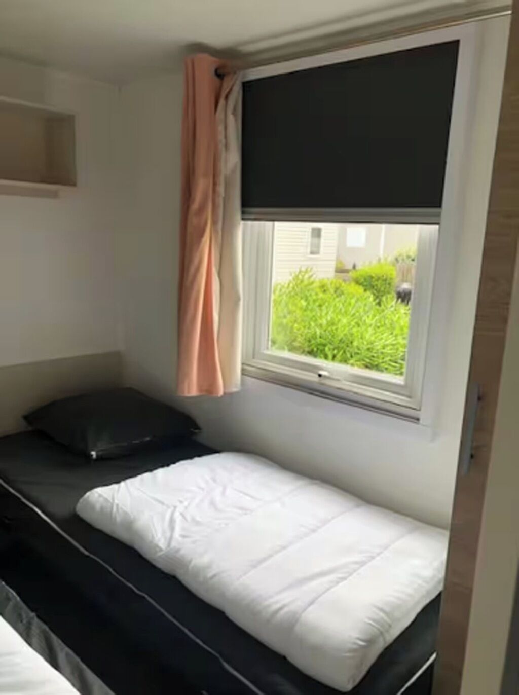 A small, single-bed room with a window, a curtain, and a view of greenery outside.