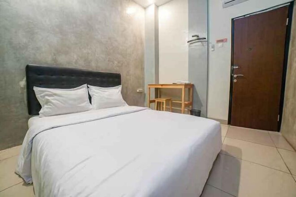 Ein Einzelbett mit Kopfteil, ein Nachttisch aus Holz und eine Tür in einem Zimmer mit schlichter Wand und gefliestem Boden.