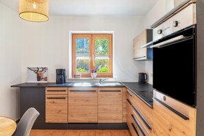 Een moderne keuken met houten kastjes, een zwart aanrecht, een raam met uitzicht op groen en een hanglamp.