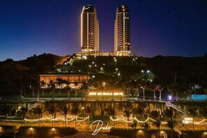 Een hotel met twee hoge torens, een goed verlichte ingang en een aangelegde omgeving met palmstruiken.