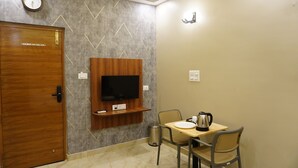 A hotel room with a wooden door, a TV mounted on a wooden shelf, a table with chairs, a kettle, and a trash bin. - Stazy Suites - Kammanhalli (Bengaluru)