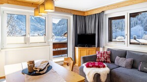 A cozy room with a wooden ceiling, a television on a wooden cabinet, a grey sofa with a white fur throw, and a dining table with a coffee cup and towels. - Apartment 'Gaichtspitze - 8' with Mountain View, Private Balcony and Wi-Fi (Nesselwängle)