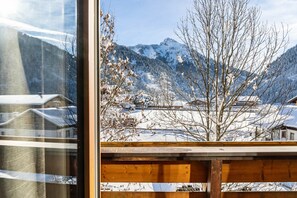A window view of a snowy mountain landscape with bare trees and buildings in the foreground.