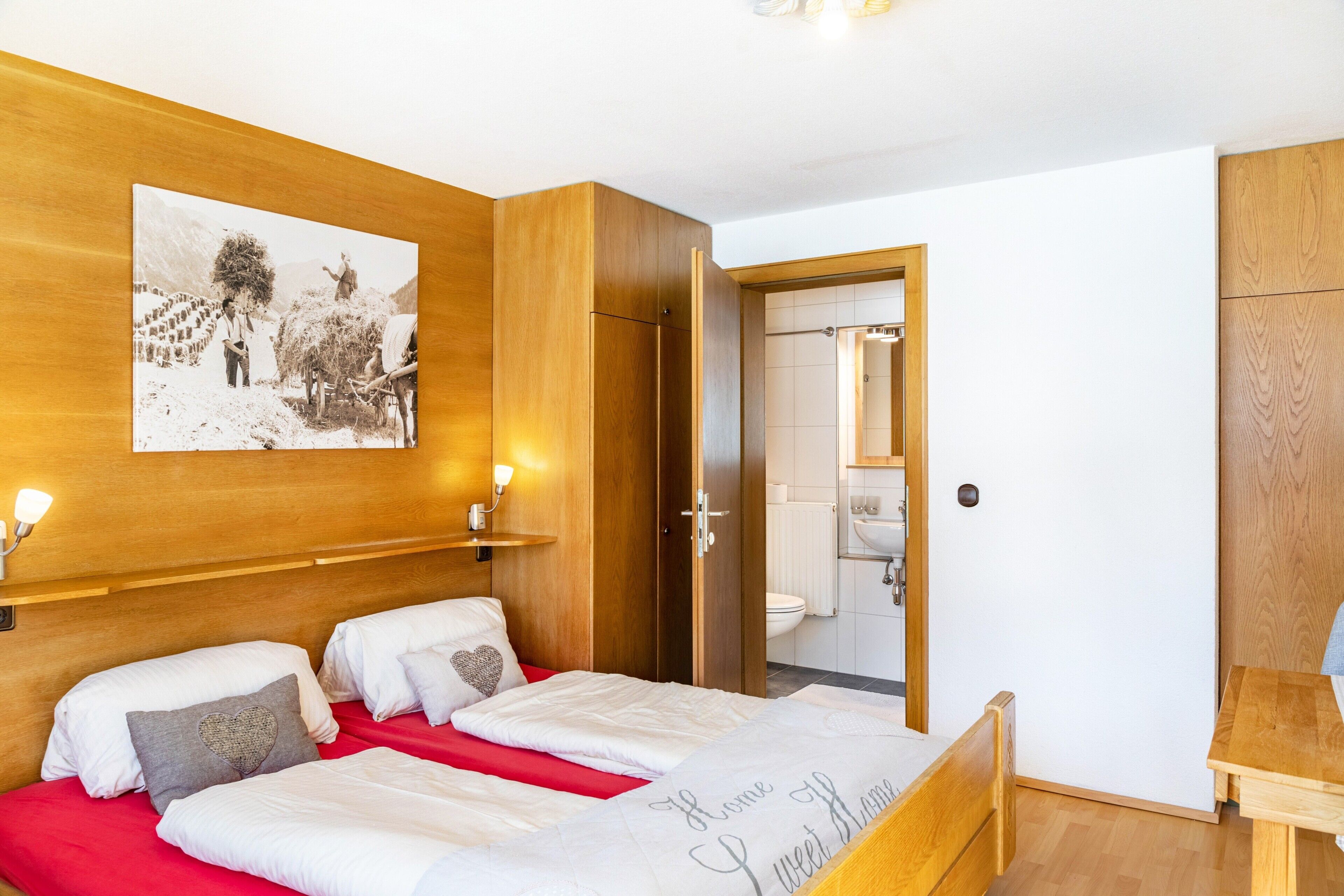 A bedroom with a wooden headboard, a bed with white bedding, a red accent, a bedside table, and a bathroom visible through an open door.