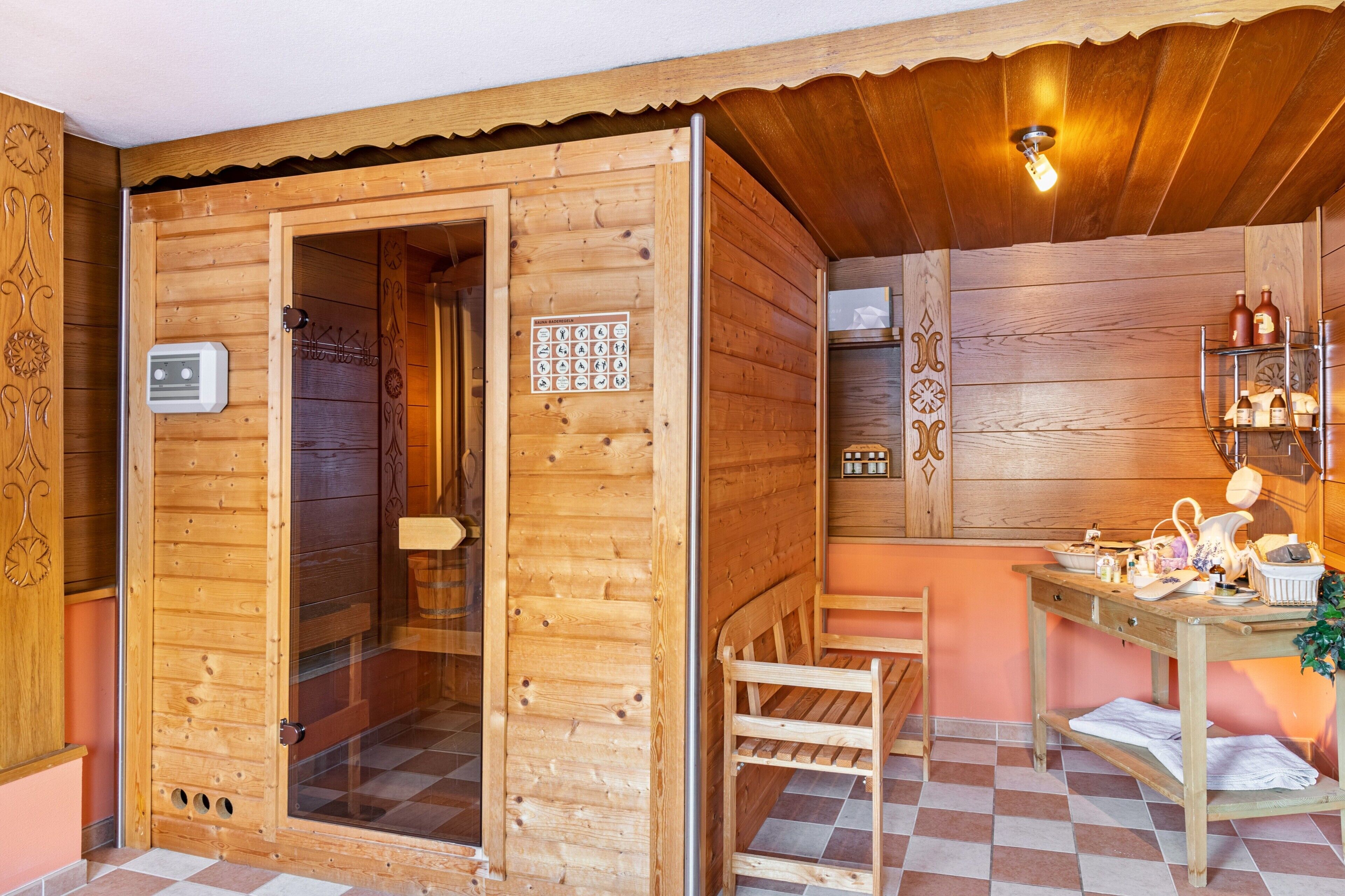 A wooden sauna room with a glass door, a bench, a table with bottles, and a wall-mounted shelf.