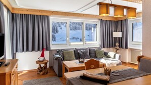 Ein gemütliches Zimmer mit einer Couch, einem Esstisch, einem Fernseher und Blick auf eine verschneite Landschaft durch große Fenster.