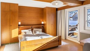 A wooden bedroom with a bed, bedside lamps, and a window with a view of a snowy landscape. - Apartment 'Hahnenkamm - 12' with Mountain View, Private Balcony and Wi-Fi (Nesselwängle)