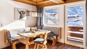 A cozy room with a wooden ceiling, a dining table with chairs, a bench, and a large window with a snowy mountain view. - Apartment 'Hahnenkamm - 12' with Mountain View, Private Balcony and Wi-Fi (Nesselwängle)