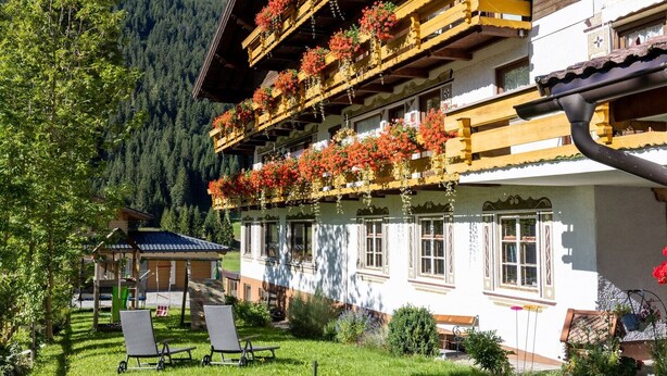 A traditional chalet with wooden balconies, a garden with lawn chairs, and a mountain backdrop. - Apartment 'Schneidspitze - 18' with Mountain View, Private Balcony and Wi-Fi (Nesselwängle)