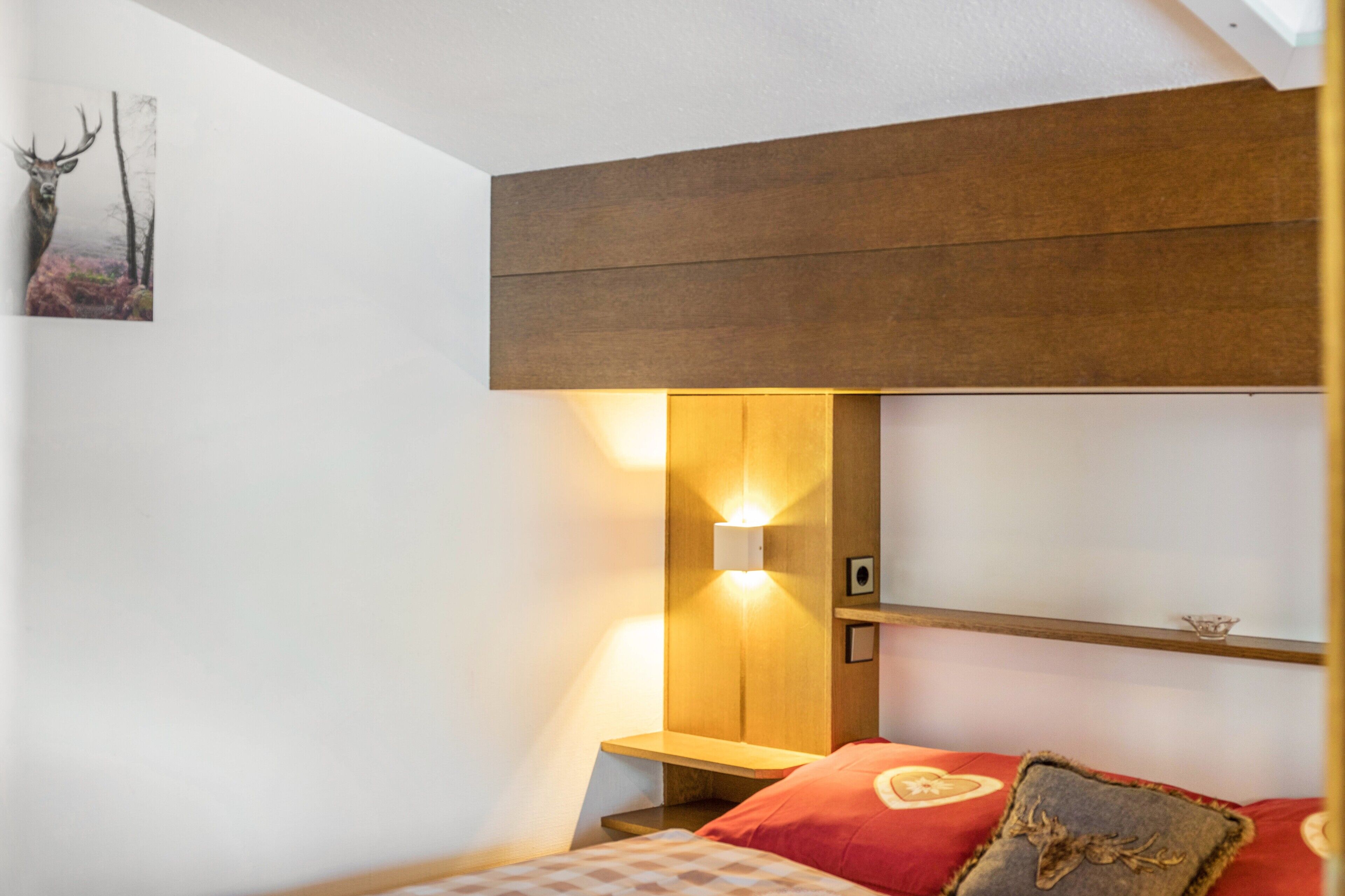 Ein Schlafzimmer mit hölzernem Kopfteil, einer Wandleuchte, einem Bilderrahmen mit einem Reh und einem roten Kopfkissen mit Herzmuster.