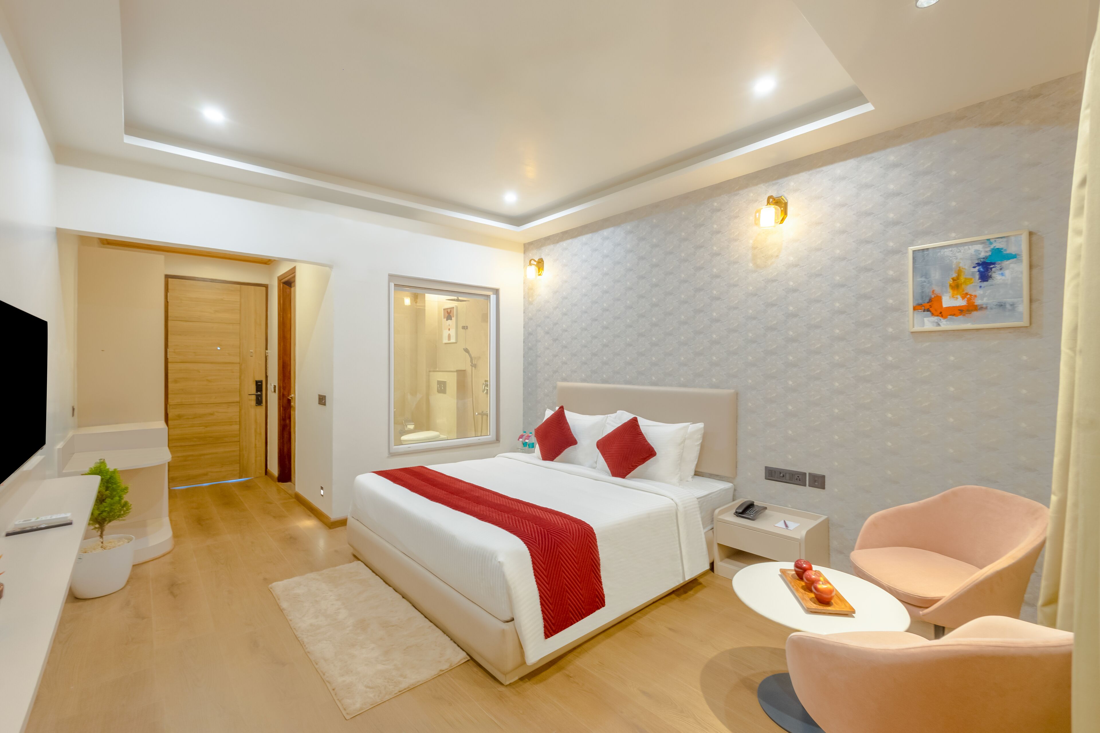 A modern hotel room with a large bed, bedside tables, a TV, a small plant, and a dining area with a table and chairs.