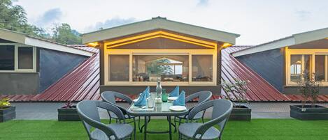 Comedor con una mesa dispuesta para cuatro personas, rodeada de sillas, frente a un edificio de diseño moderno y amplias ventanas.