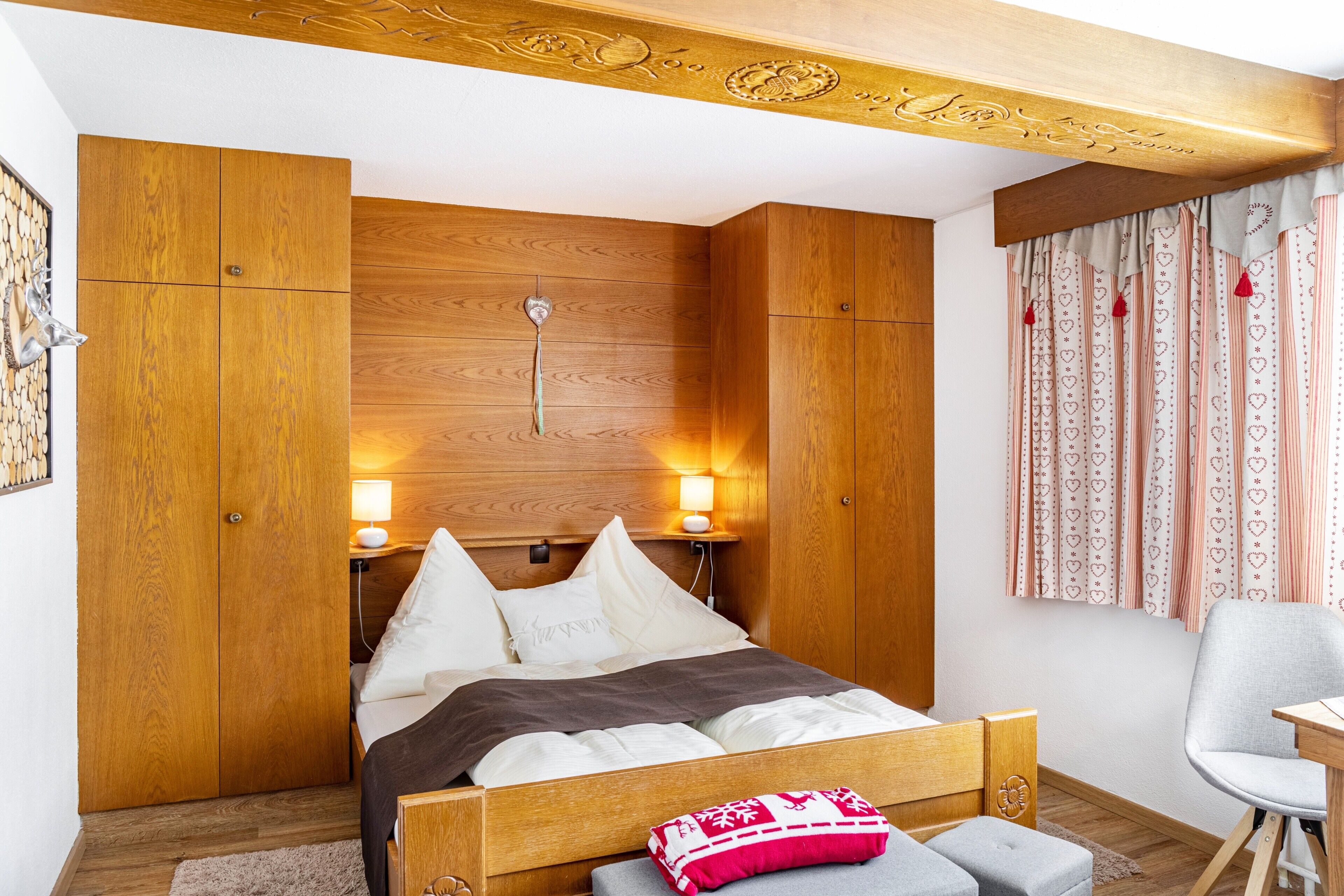 Een slaapkamer met een houten hoofdbord, een bed met wit en bruin beddengoed, twee bedlampen en een raam met patroon gordijnen.