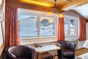 Ein Zimmer mit Tisch, zwei Stühlen, einer Lampe und Blick auf eine verschneite Landschaft durch große Fenster.