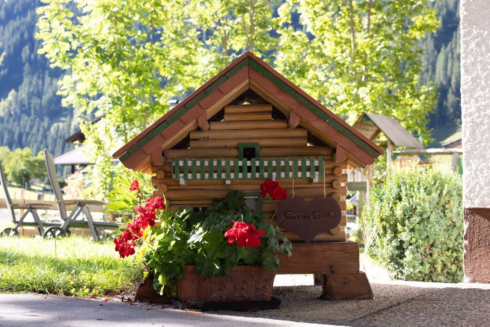 Un comedero de pájaros de madera con una placa en forma de corazón que dice 'Griss Gott' rodeado de flores rojas.